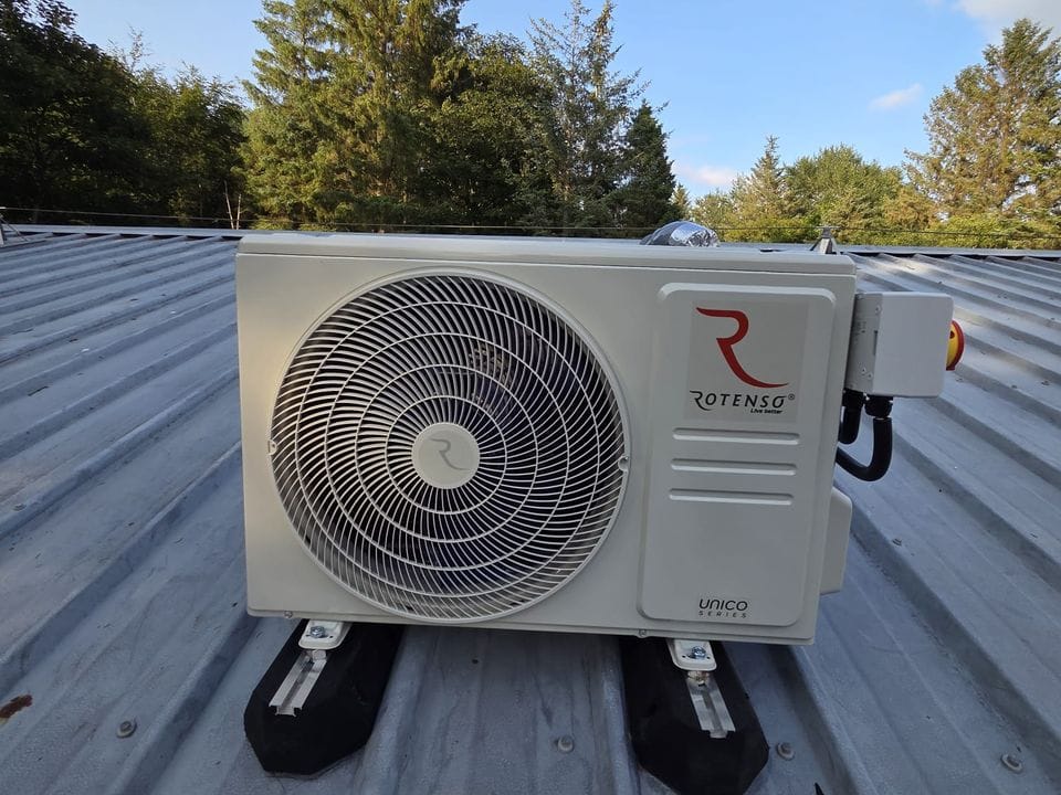Rotenso outdoor unit standing on raised feet on a corrugated metal roof, with conifers behind